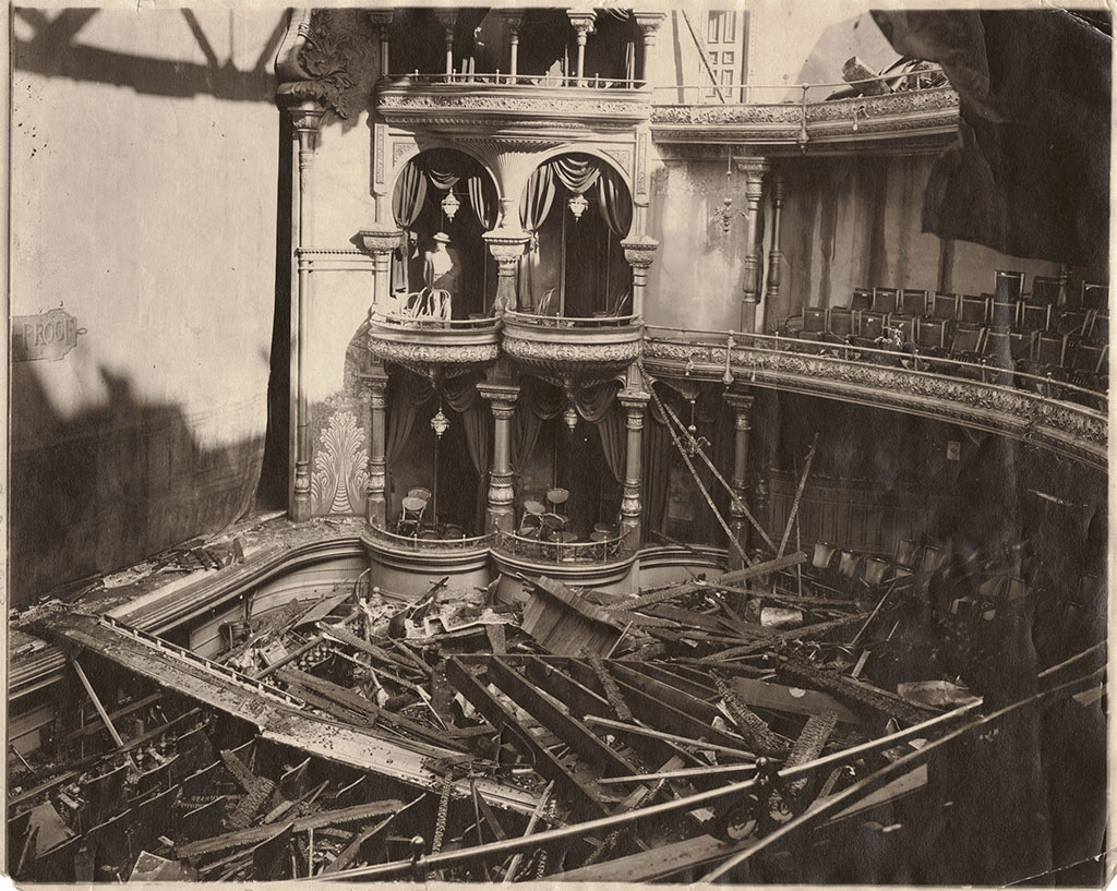 Illustration from the novel theMystery.doc by Matthew McIntosh. Historic sepia-toned photograph of a man standing on a theatre balcony overlooking what was once the seating area. The theatre roof has collapsed, and lumber and beams cover the rows of seats. The stage curtain is down, and on the curtain is the word "Proof".