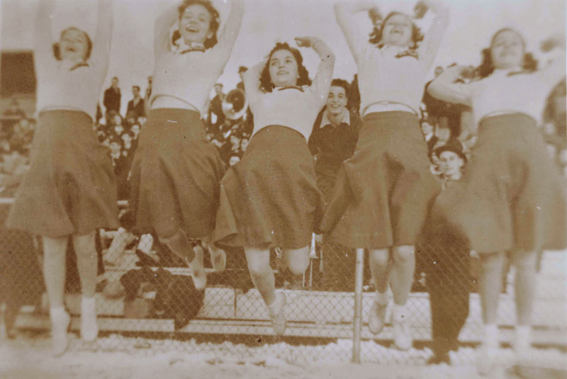 Illustration from the novel theMystery.doc by Matthew McIntosh. Sepia-toned photograph of a row of five smiling female cheerleaders jumping in the air. They are wearing long-sleeved sweaters, knee-length skirts, and bobby socks. A young man in a dark suit and fedora looks out from behind them.