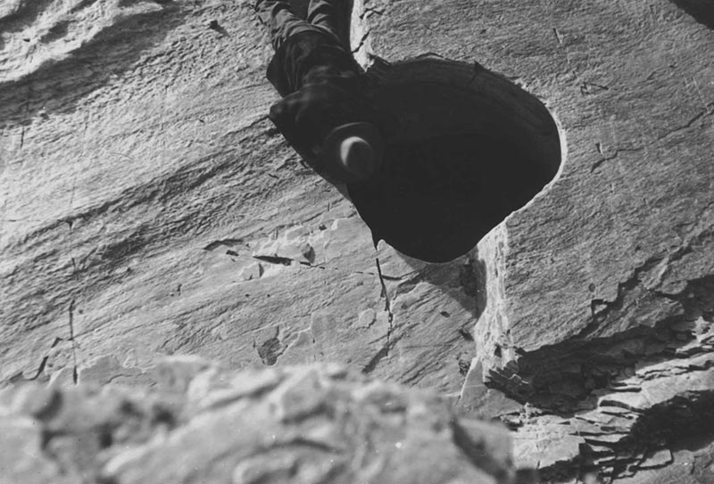 Illustration from the novel theMystery.doc by Matthew McIntosh. Black-and-white photograph of a man looking down a dark shaft in a rocky ledge.